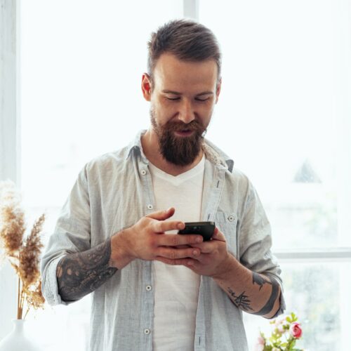 Handsome young bearded man spending time at home with phone in hands.